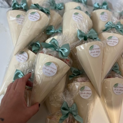 Cones de papel creme embalados em celofane com laços verdes e etiquetas redondas com texto sobre batizado.