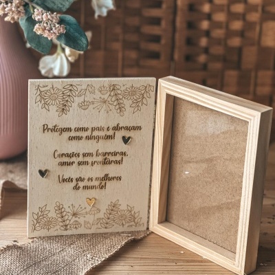 Moldura dupla de madeira clara gravada com texto e flores e corações decorativos, em cima de madeira e tecido, com jarro e flores ao fundo