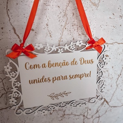 Placa decorativa branca com texto dourado pendurada por fita vermelha com laços
