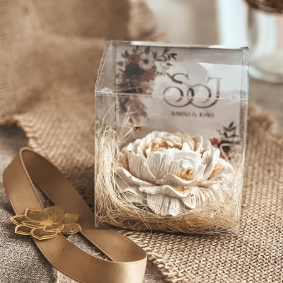 Flor branca decorativa em caixa transparente com laço dourado sobre juta