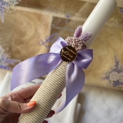Vela branca decorada com corda branca, laço roxo, flores secas lilás e placa dourada com texto gravado