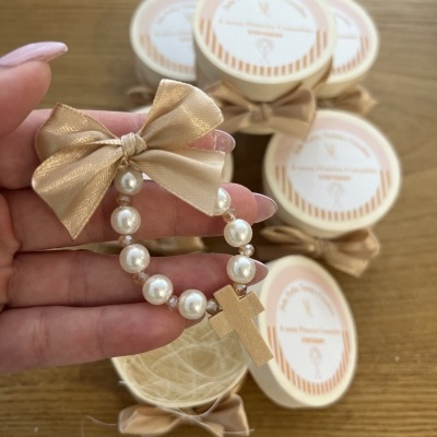 Pulseira de pérolas com laço dourado e cruz dourada, com pequenas caixas redondas brancas com laços e etiquetas em português sobre uma mesa de madeira.