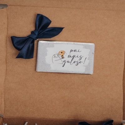Caixa de cartão castanha aberta com laço azul e barra de chocolate com texto em papel branco