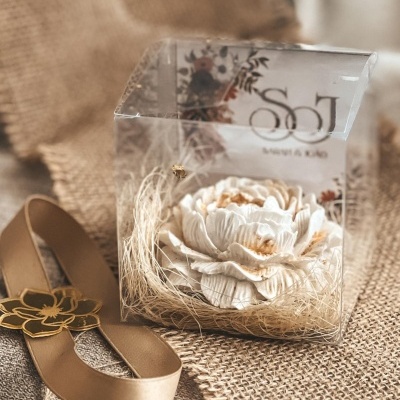 Flor decorativa branca numa caixa transparente com palha e fita bege com flor dourada