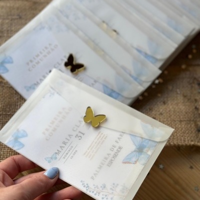 Convite de primeira comunhão com envelope translúcido e pin de borboleta dourado.