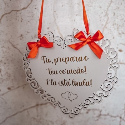 Placa de coração branca com texto dourado e laços vermelhos pendurada numa parede clara