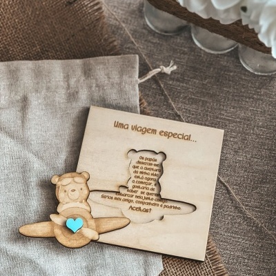 Quadro de madeira com avião e urso segurando coração azul sobre tecido cinza e juta com flores brancas ao fundo