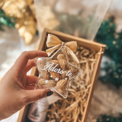 Ornamento estrela transparente com amendoins e texto Adoro-te, laço dourado, mão segurando, caixa presente ao fundo
