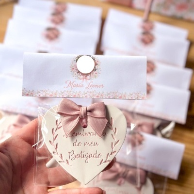 Lembrança de batizado em forma de coração com laço rosa e cartão floral branco com texto.