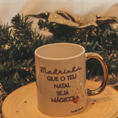 Caneca branca com texto natalício e asa dourada sobre base de madeira com decoração de Natal