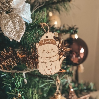 Enfeite de Natal gato de madeira com cachecol e gorro escrito Evy numa árvore verde decorada com bolas e folha dourada.