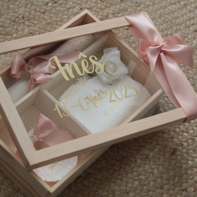 Caixa de madeira com tampa de vidro e laço rosa contendo um objeto branco com inscrição dourada e flor branca
