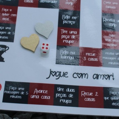 Tabuleiro de jogo branco com textos em português, dado branco com pontos vermelhos e peças em forma de coração