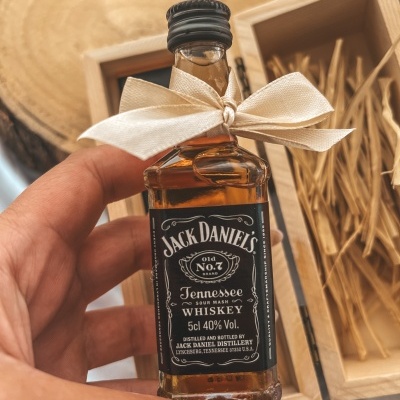 Frasco pequeno de whiskey Jack Daniel's com laço bege, sobre caixa de madeira