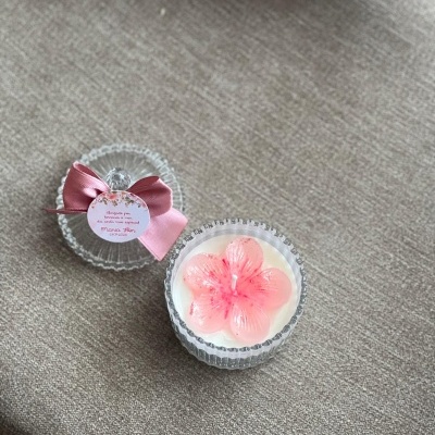 Vela branca em recipiente de vidro com tampa e laço rosa numa superfície cinza