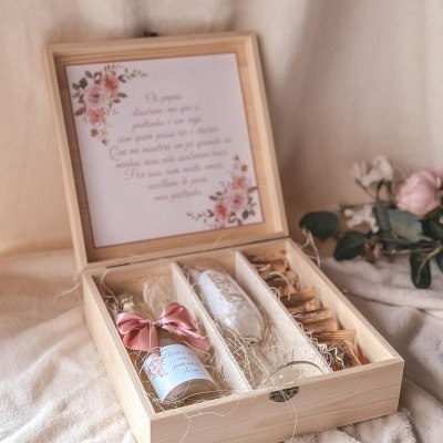 Caixa de madeira clara com garrafa, caneca e pacotes dentro, junto a um cartão com texto em Português e decoração floral.