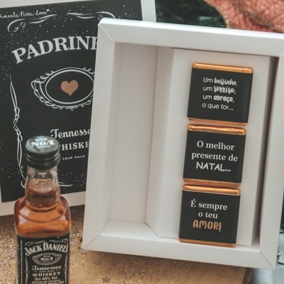 Mini garrafa de whisky Jack Daniel's ao lado de caixa branca com três chocolates pretos com texto em português, com embalagem preta decorativa ao fundo