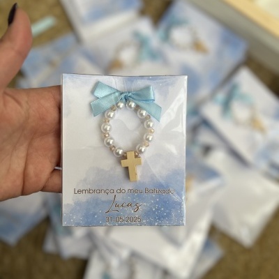 Pulseira branca com cruz dourada e laço azul em cartão de batizado com texto