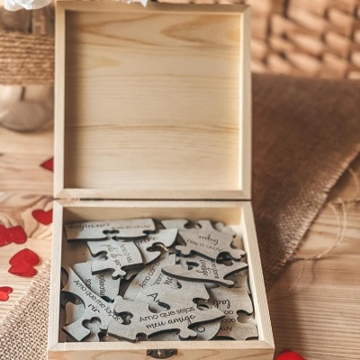 Caixa de madeira aberta com peças de puzzle cinzentas com texto em português, sobre mesa de madeira com detalhes em tecido e corações vermelhos.