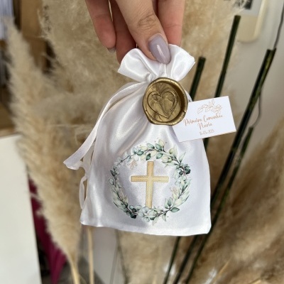 Saco branco de cetim com cruz dourada e coroa floral, selo de cera dourado e etiqueta branca, mãos de uma pessoa segurando