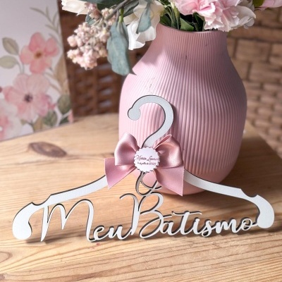 Cabide branco decorativo com laço rosa e texto Meu Batismo junto a vaso rosa com flores artificiais