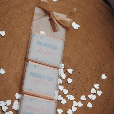 Três chocolates embrulhados com texto em papel branco, colocados sobre base de madeira, com confetes em forma de coração branco.