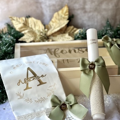 Conjunto de batismo com toalha branca bordada, vela decorada com laço verde e concha com laço, colocados sobre superfície branca e caixa ao fundo.