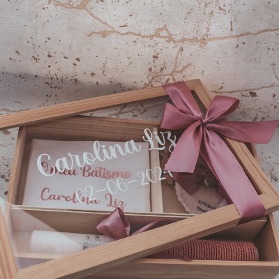 Caixa de madeira com tampa de vidro e laço rosa com velas, toalha e cartão de batismo