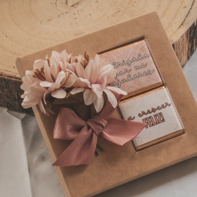 Caixa de presente com flores artificiais e dois chocolates com texto impresso, sobre superfície branca e suporte de madeira