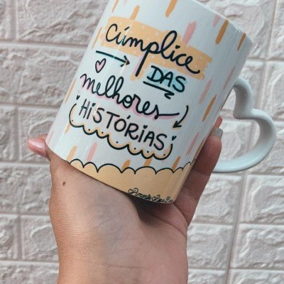 Caneca branca com alça em forma de coração e texto decorativo, segurada por mão com tatuagem, parede de tijolos rosa ao fundo