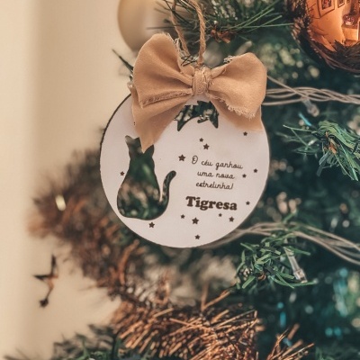 Ornamento redondo branco com texto e recorte de gato numa árvore de Natal verde.