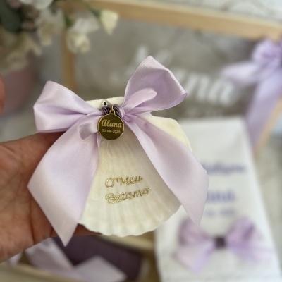 Concha branca com laço lilás e medalha dourada com nome e data, inscrições douradas, decoração de batismo