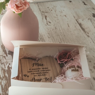 Caixa branca com presente para mãe, bloco de madeira com mensagem, vela decorada e flor, ao lado de vaso rosa com flores