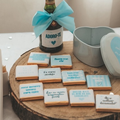 Garrafa com laço azul claro e chocolates com mensagens em embalagem branca e azul numa base de madeira
