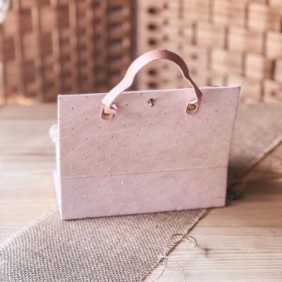 Mala rosa clara texturizada com pequenos detalhes metálicos e alça rosa sobre mesa de madeira e tecido bege.