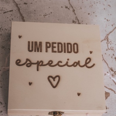caixa de madeira clara com gravação 'UM PEDIDO especial' e corações em fecho metálico sobre superfície bege com manchas