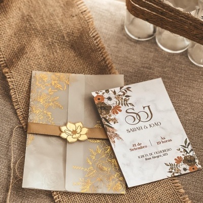 Convite de casamento com cartão floral e envelope translúcido com detalhes dourados em fundo bege rústico