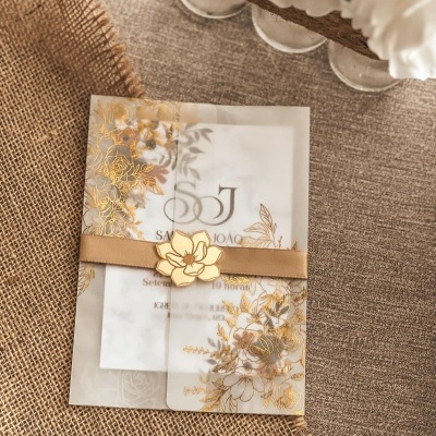 Convite translúcido com detalhes dourados e laço bege sobre tecido rústico