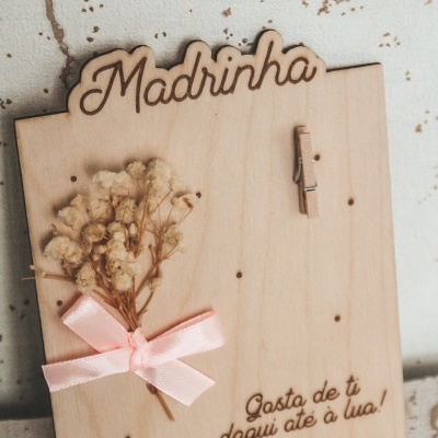 Placa decorativa de madeira com texto gravado, flores secas e fita rosa.
