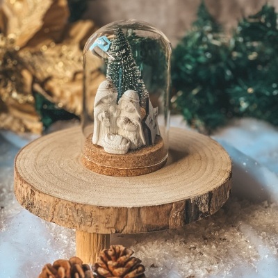 Presépio miniatura em campânula de vidro sobre suporte de madeira com pinhas e neve artificial