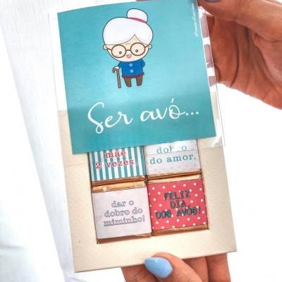 Caixa de chocolates com mensagens de feliz dia dos avós segurada por mãos femininas com unhas azuis.