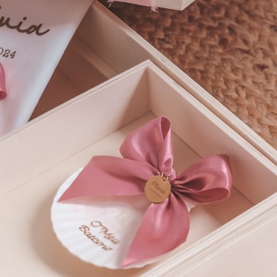 Concha branca com fita rosa e medalha dourada dentro de caixa branca.