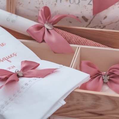Conjunto para celebração com pano branco bordado, velas e laços rosa numa caixa de madeira