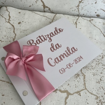 Livro de assinaturas para batizado com capa branca, texto rosa e laço rosa sobre superfície manchada