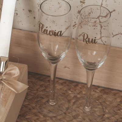 Dois copos de vidro com nomes escritos em dourado, caixas de madeira com fitas bege e laços, em superfície de tecido castanho.