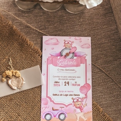 Convite de batizado rosa com desenhos infantis, texto e detalhes decorativos sobre tecido castanho