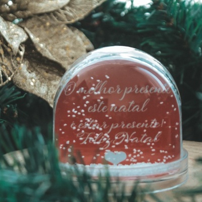 Globo de neve vermelho com mensagem de Natal rodeado de ramos de pinheiro e flor dourada