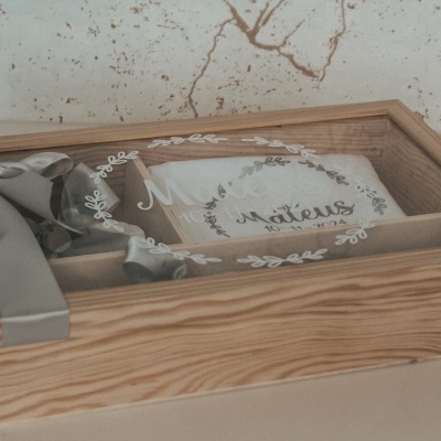 Caixa de madeira clara com laço cinzento e toalha branca bordada com floral e texto