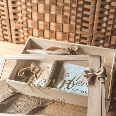 Caixa de madeira com tampa de vidro e nome Kevin dourado, com tecido branco e laços dentro, decoração em juta e fundo rústico.