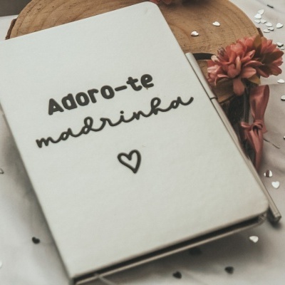 agenda branca com texto 'Adoro-te madrinha' e coração, com flores cor de rosa e confetes ao redor